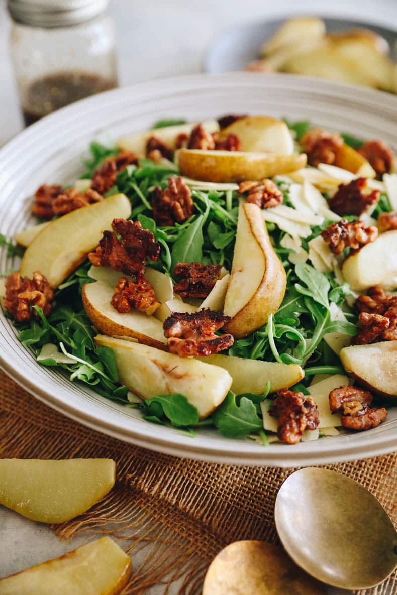 Pear salad over a bed of arugula with candied walnuts and shaved parmesan with a balsamic vinaigrette in a large white salad bowl.