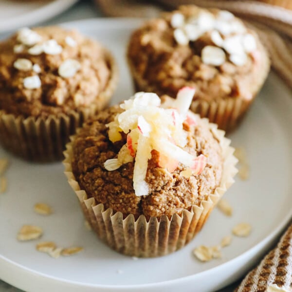 close up image of an oatmeal apple muffin topped with rolled oats and shredded apple on a white plate.