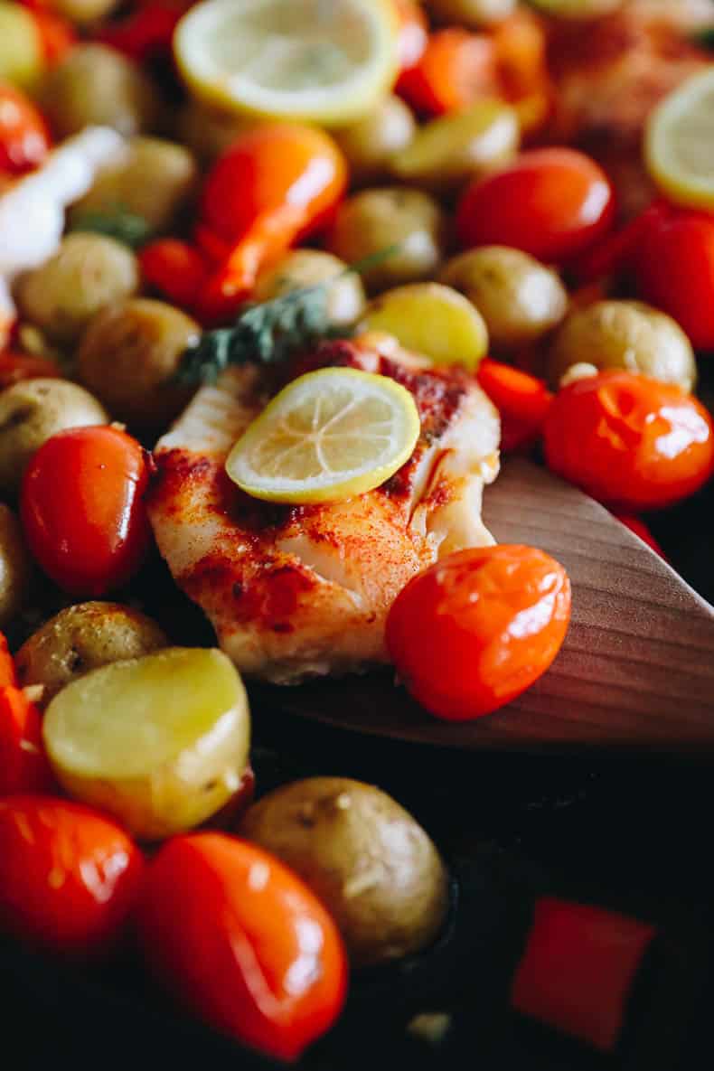 up close image of baked cod on a sheet pan with potatoes, peppers and cherry tomatoes.