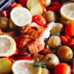 flaky baked cod with paprika on a sheet pan with lemon slices, potatoes, tomatoes and bell peppers.