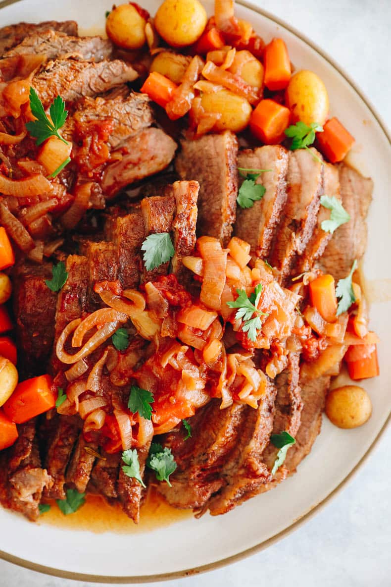 sliced brisket on a white platter with gravy, potatoes, carrots and parsley