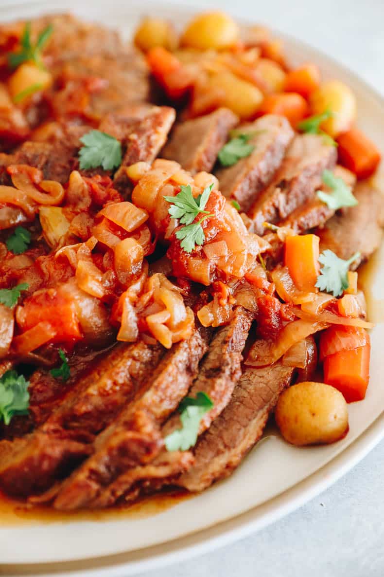 brisket recipe on a white platter with onions, carrots, potatoes and parsley