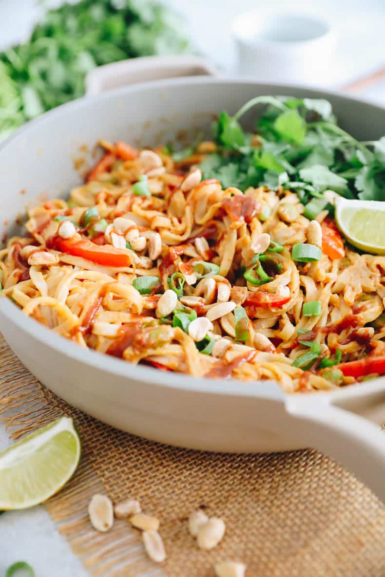 peanut noodles in a pan with scallions, sriracha, lime wedges and crushed peanuts